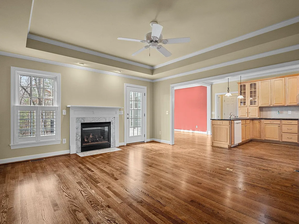 Open living room with a tray ceiling, white fireplace, warm wood flooring, and views into the kitchen