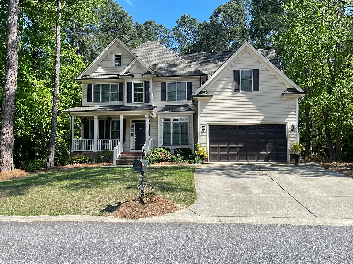 Light two-story home with layered gables, dark garage doors, a covered porch, and mature trees