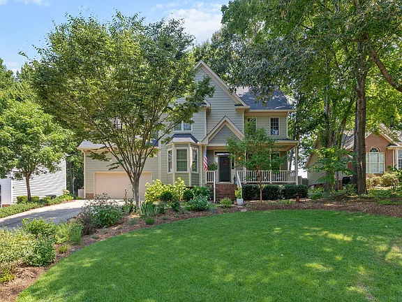 Traditional two-story home with green siding, a covered porch, mature trees, and a broad landscaped lawn