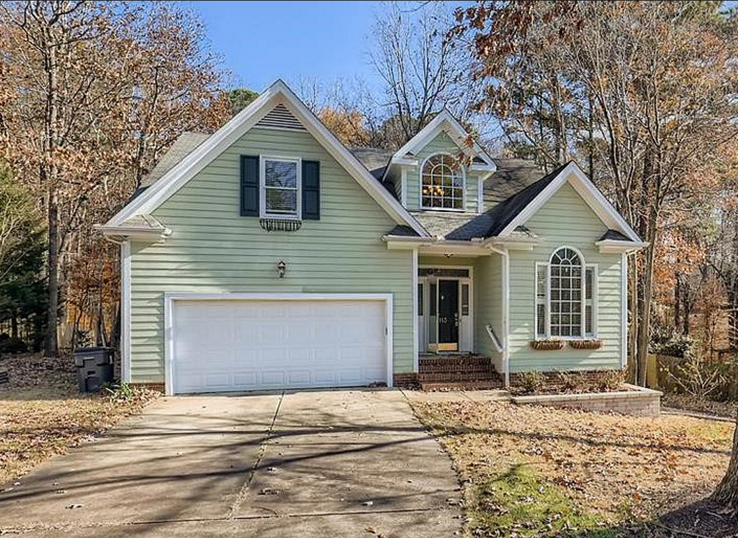 Sage green home with layered gables, arched windows, an attached garage, and a wooded setting