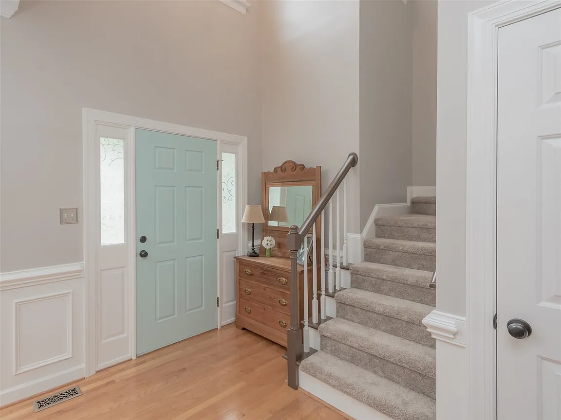 Bright entry hall with a pale blue front door, warm wood flooring, and a carpeted staircase