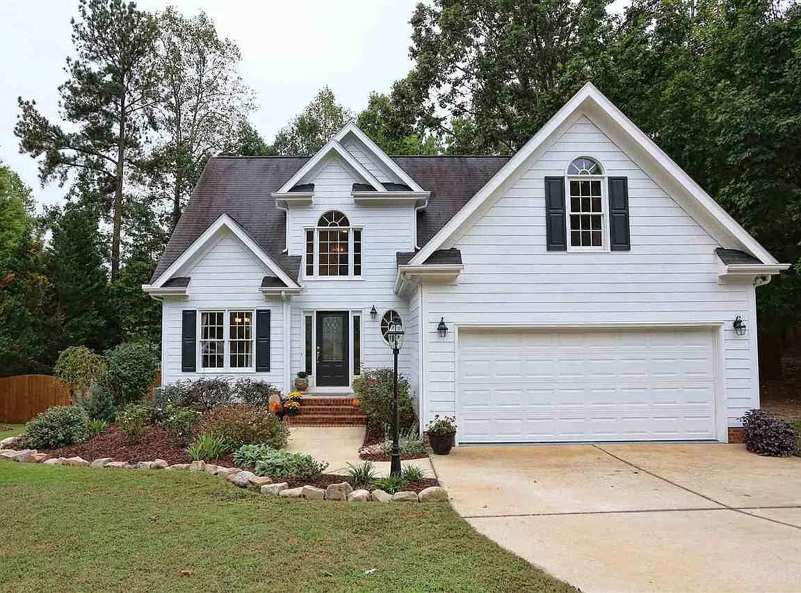 White two-story home with layered gables, dark shutters, an attached garage, and a landscaped front yard