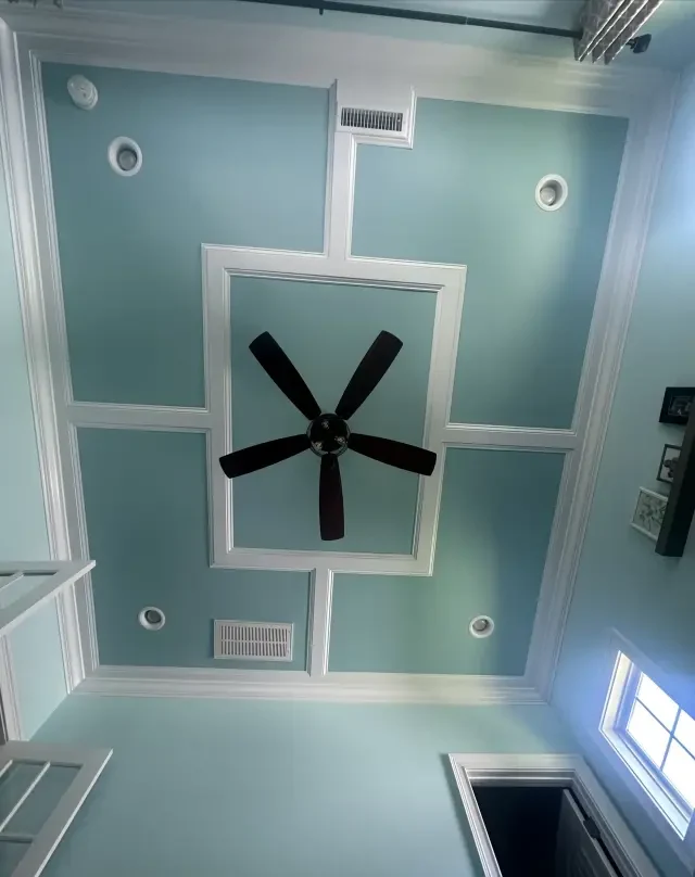 Blue bedroom ceiling framed by white geometric millwork around a central ceiling fan